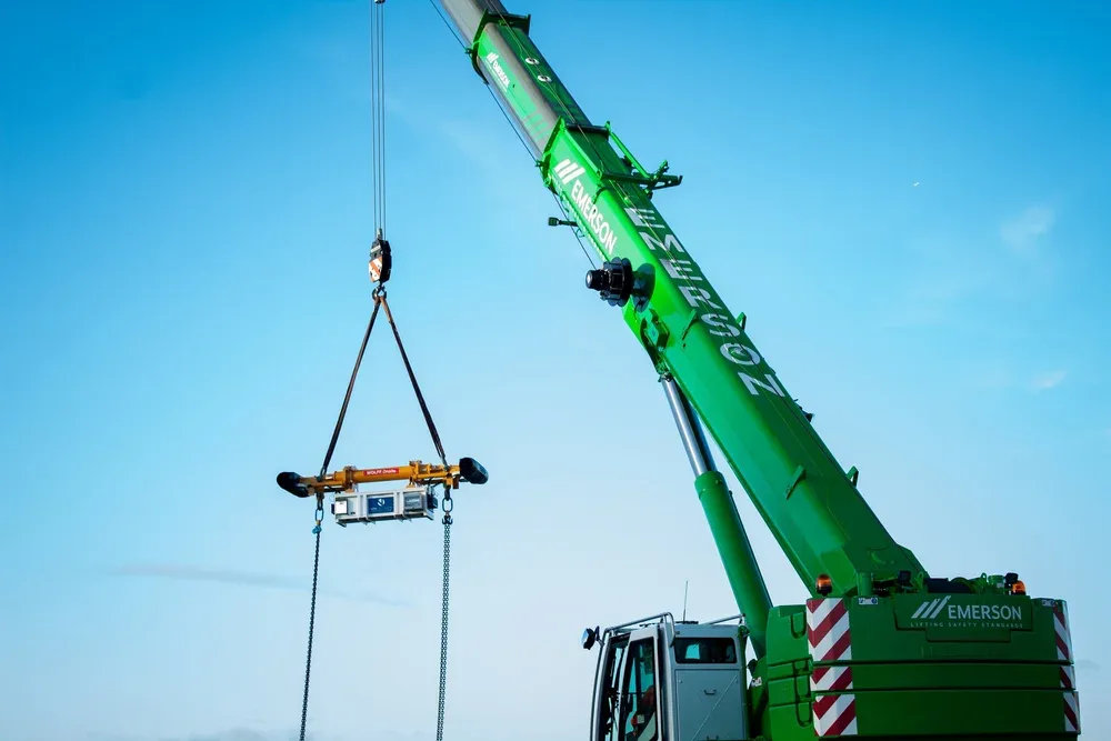 Green crane lifting a piece of equipment with chains against a blue sky.