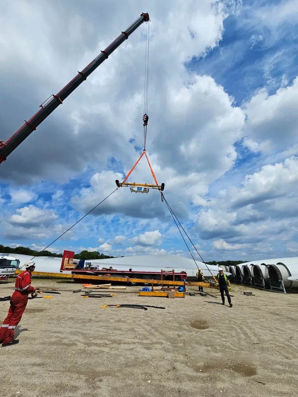Construction workers operate a crane lifting a large piece of equipment outdoors with wind turbines in the background and a partly cloudy sky.