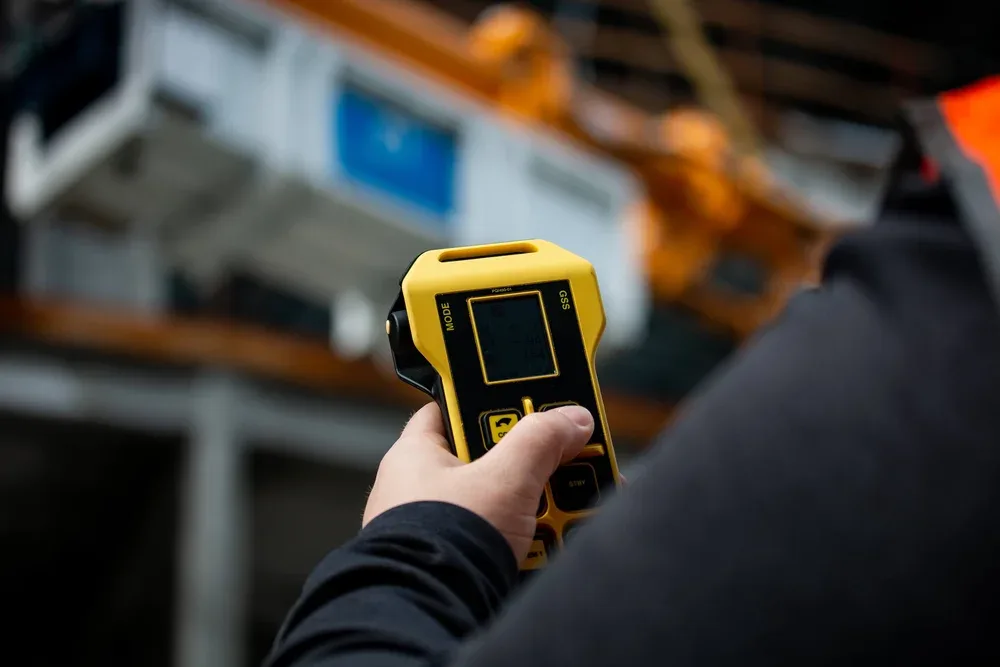 Person holding a digital measuring device in an industrial setting with blurred machinery and structures in the background.