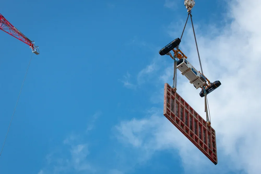A construction crane lifting a large red and black building panel against a blue sky with some clouds.