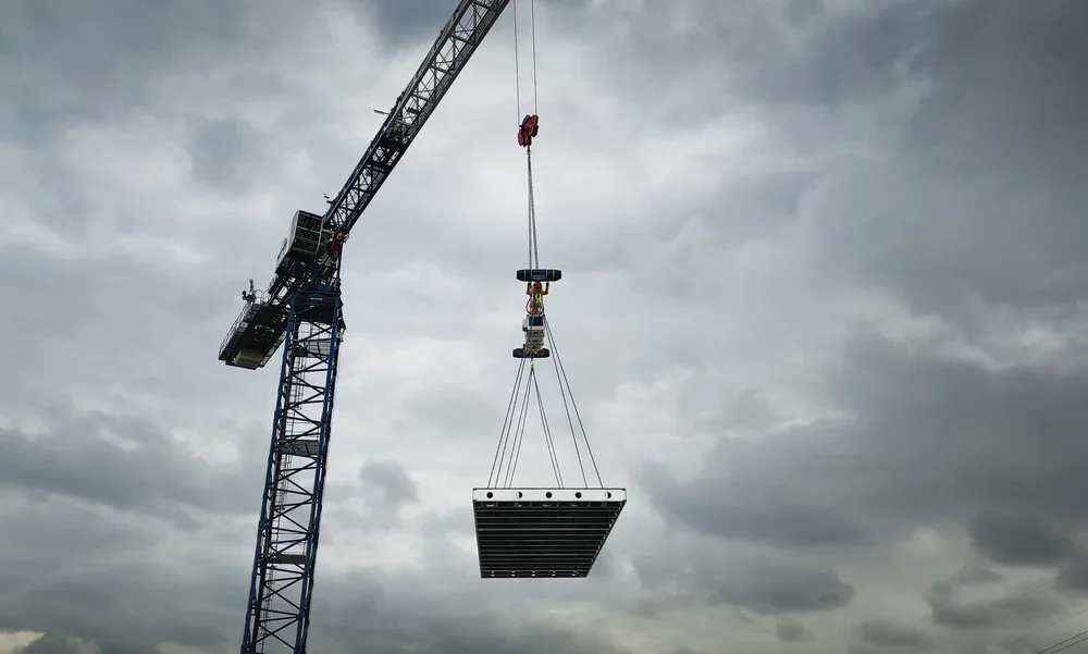 A construction crane lifting a large metal platform against a cloudy sky.