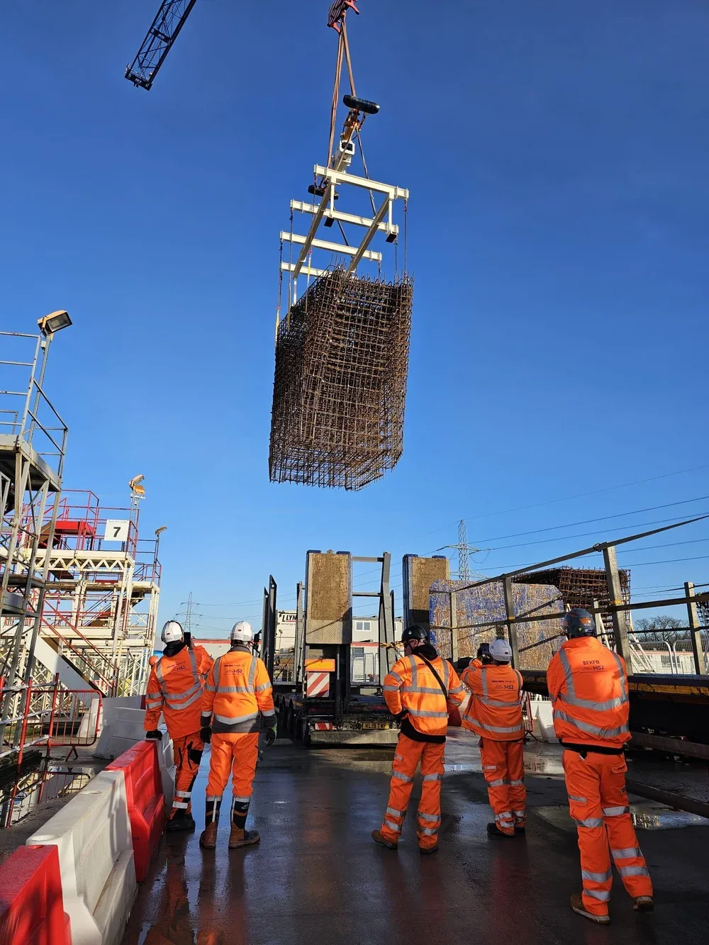 Construction site with workers in orange safety gear and helmets watching a large steel rebar structure hanging from a crane against a clear blue sky.