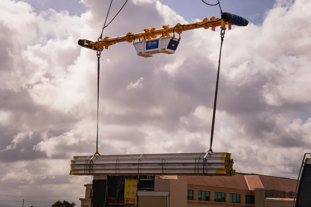 Construction crane lifting long metal beams above a building with a cloudy sky in the background.