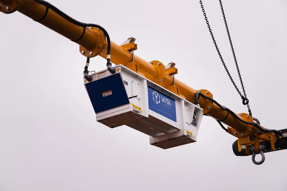 A large industrial crane with a yellow boom and a blue control box labeled 'VITA INDUSTRIAL LOAD NAVIGATOR' suspended in the air against a gray overcast sky.