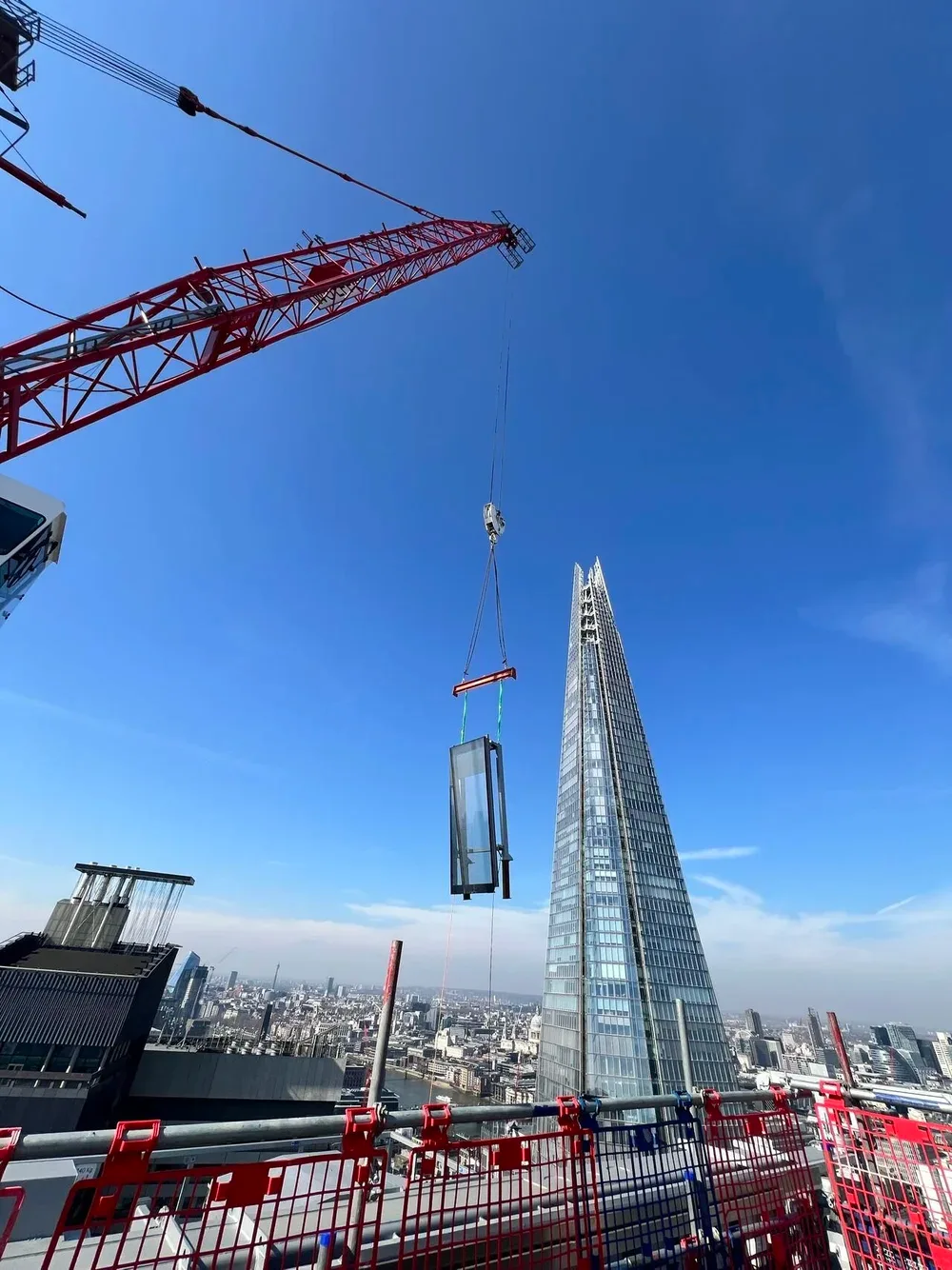 A construction site on a high-rise building in London with a crane lifting a glass panel, the Shard in the background, and a city skyline under a clear blue sky.