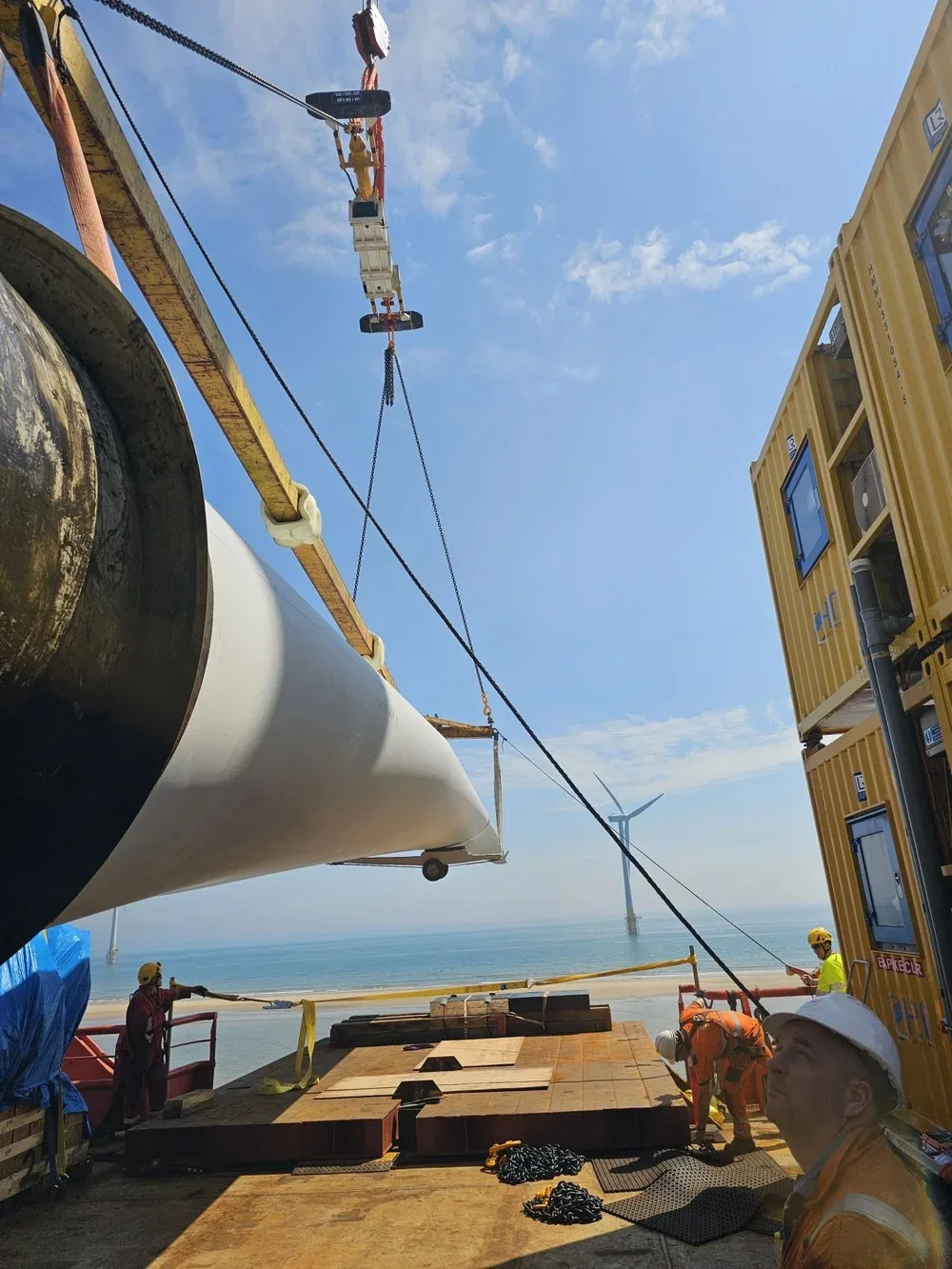 Construction workers in safety gear work on installing a large wind turbine blade at a seaside offshore wind farm, with wind turbines visible in the background under a blue sky.