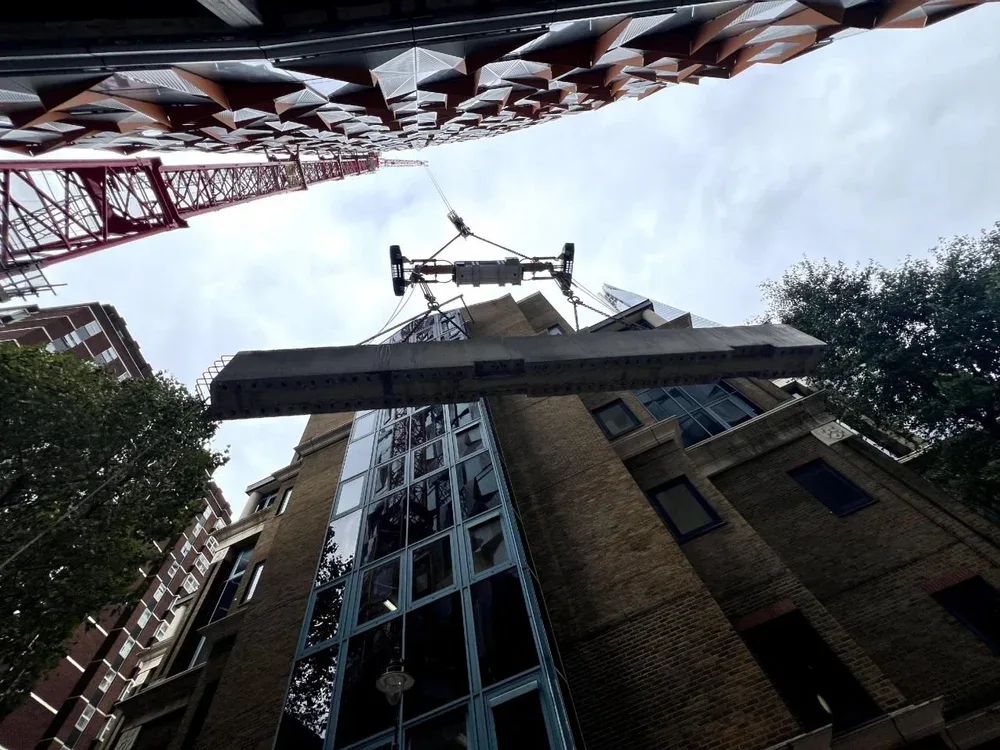 View looking up at construction crane lifting a large building panel, surrounded by tall buildings with brick and glass exteriors, and trees on the side, under a cloudy sky.
