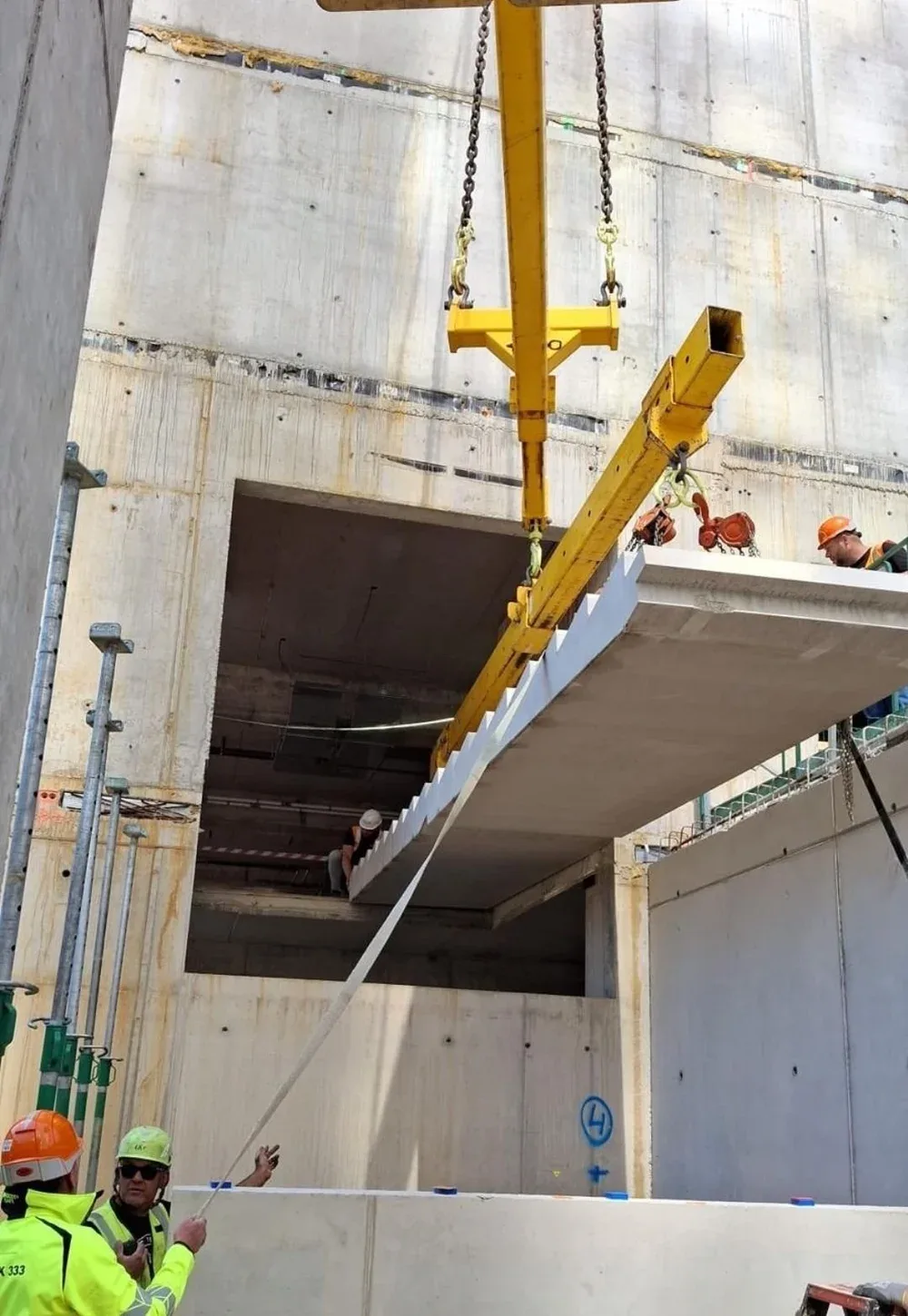 Construction workers are installing a prefabricated concrete section with the help of a yellow lifting crane at a building site.