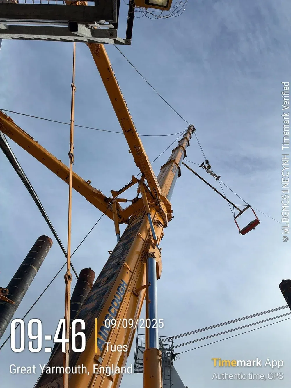 A construction crane with a yellow arm and a white boom, extending into a sky with some clouds. The crane has a platform with a red safety net at the end of the extended arm. There are black cylindrical structures nearby, and electrical wires are vis