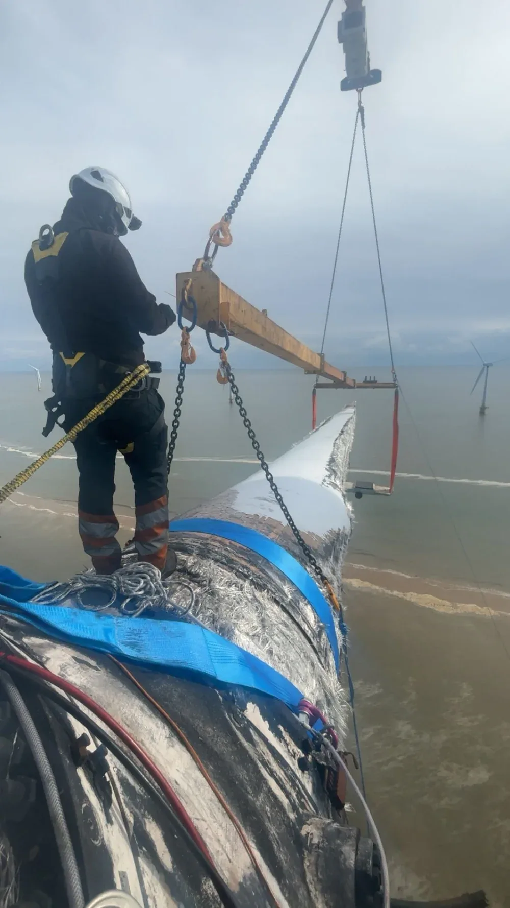 A worker in protective gear and helmet operating a crane to lift a large underwater wind turbine blade in the ocean near wind turbines.