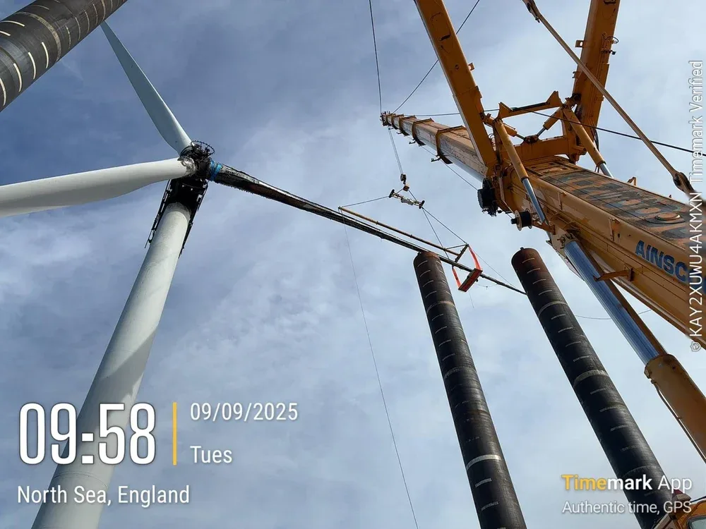 Close-up view of a crane lifting wind turbine components during construction, with a blue sky and some clouds in the background.