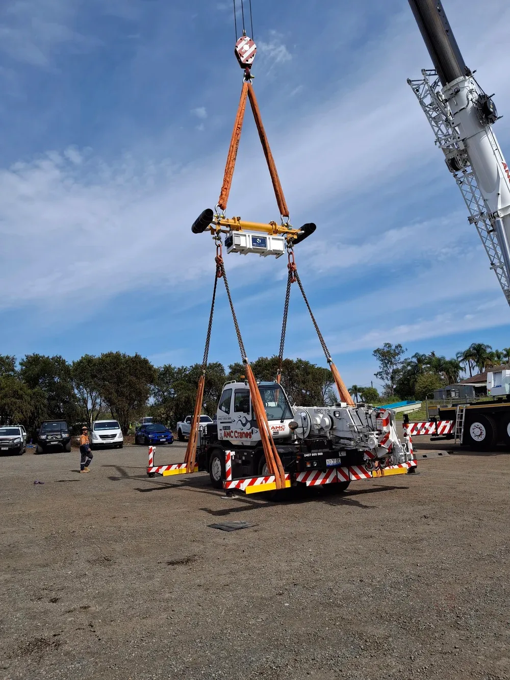 A crane suspending a large industrial machine in a parking lot with several parked cars and a person walking nearby.