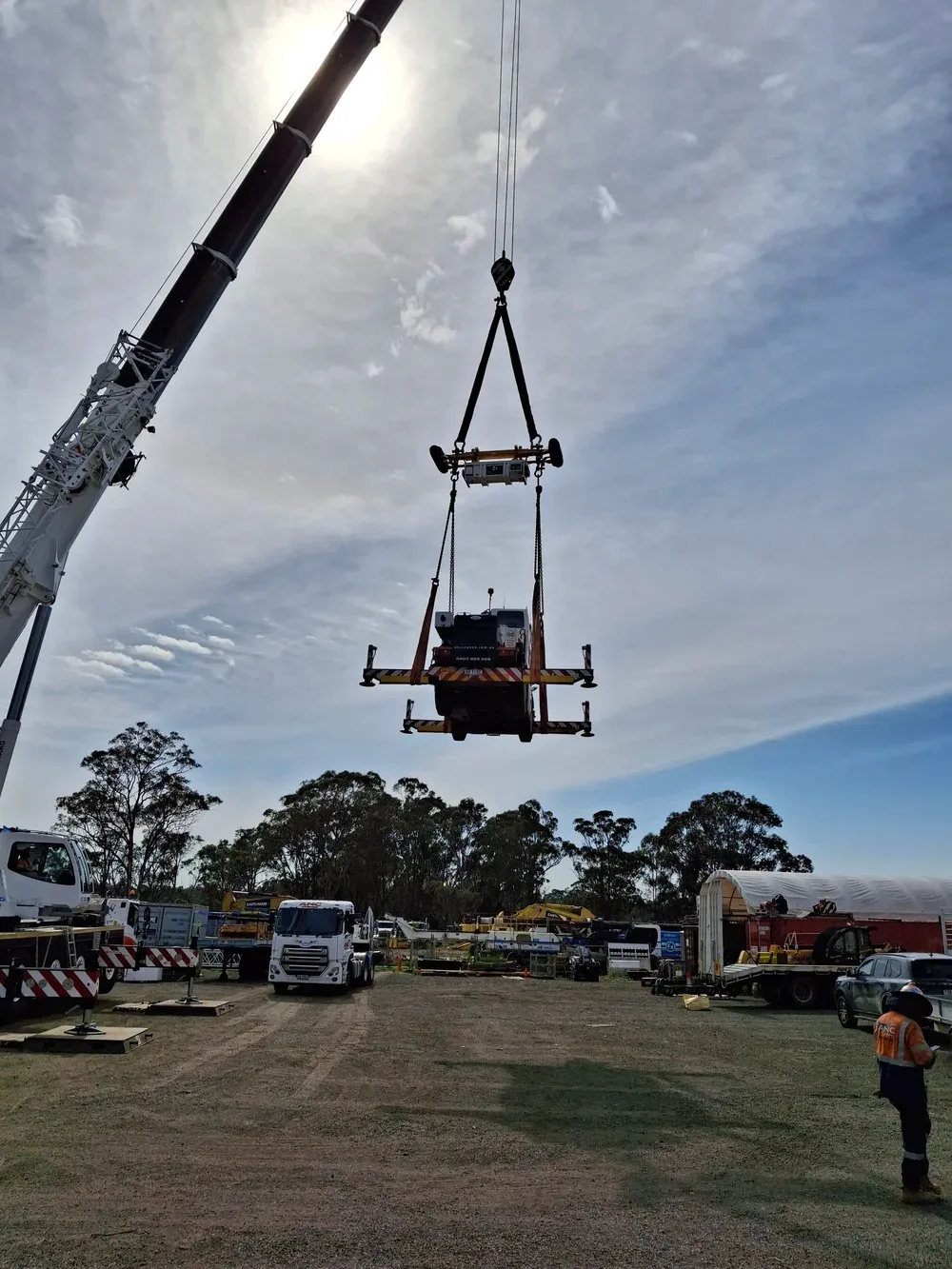 A construction crane lifting a large piece of equipment at a construction site with trucks and machinery, trees in the background, and a worker wearing an orange safety vest on the ground.