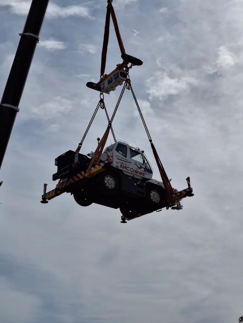 A crane lifting a small truck with the logo 'ANC Cranes' on it, against a cloudy sky.