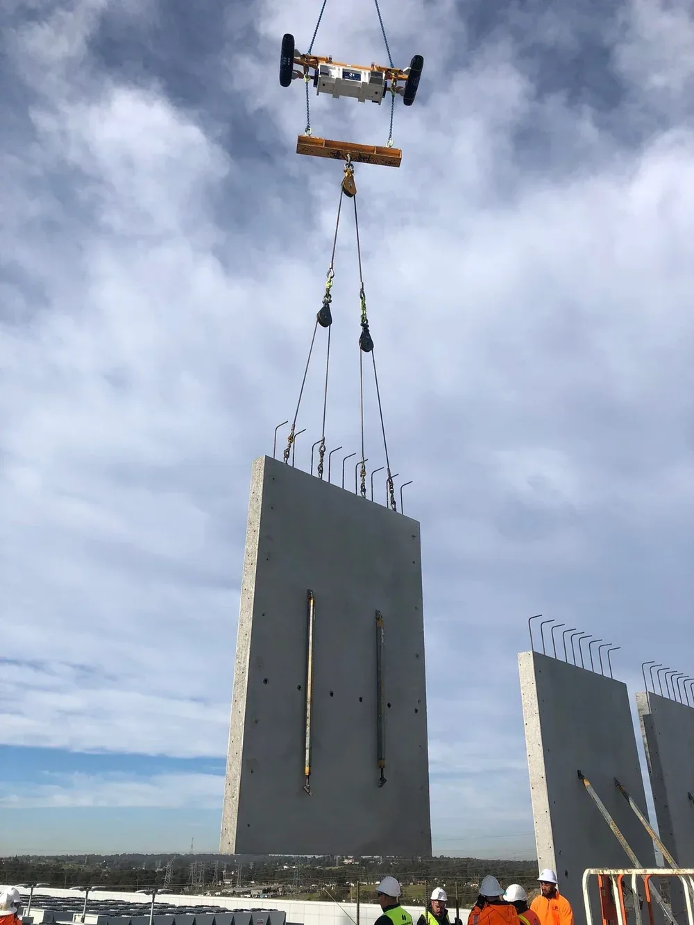 Construction workers in safety gear observe as a large concrete panel is lifted by a crane at a construction site under a cloudy sky.