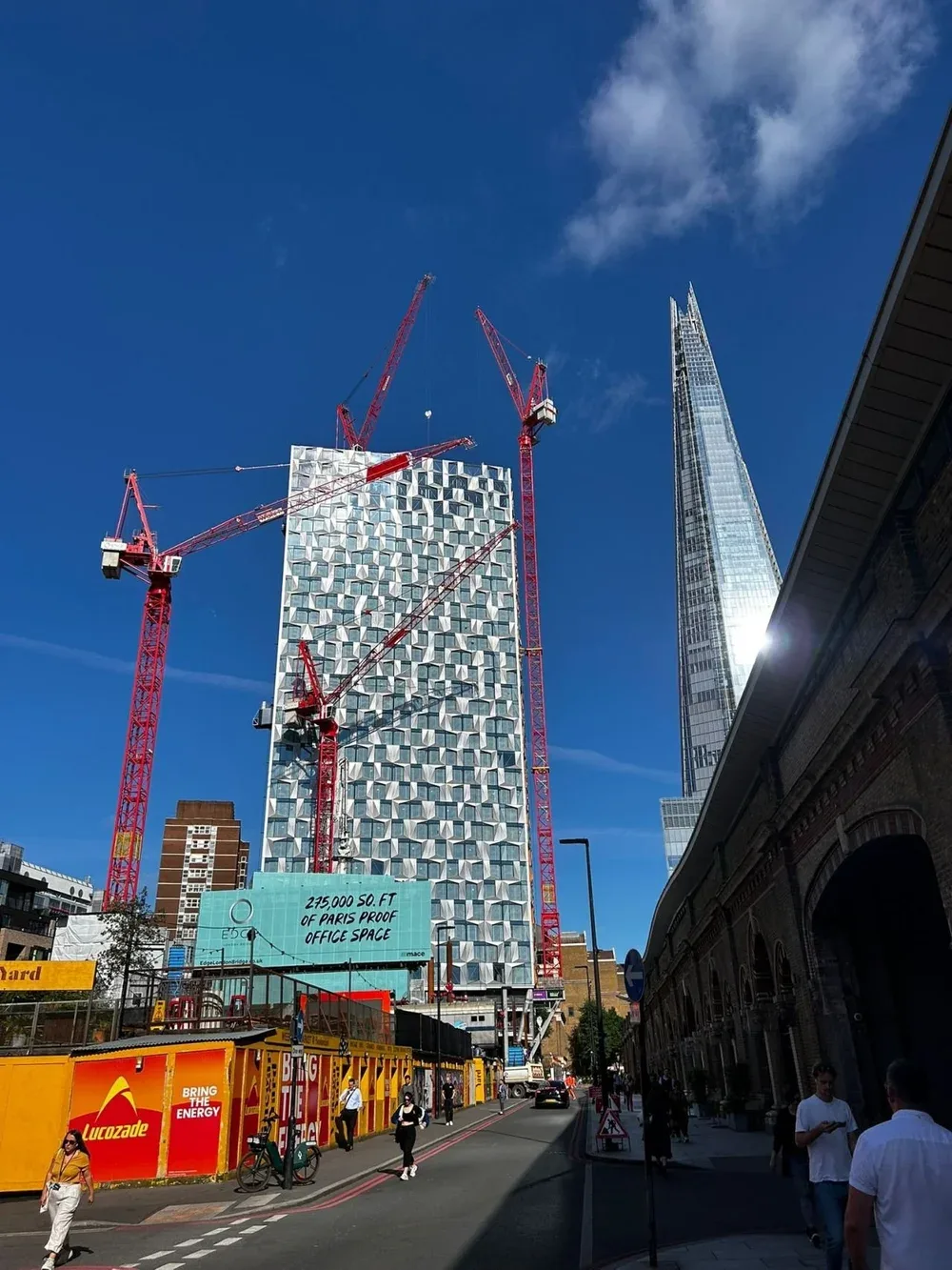 Construction site on a city street with a modern skyscraper under construction, cranes, and pedestrians walking on the sidewalk.