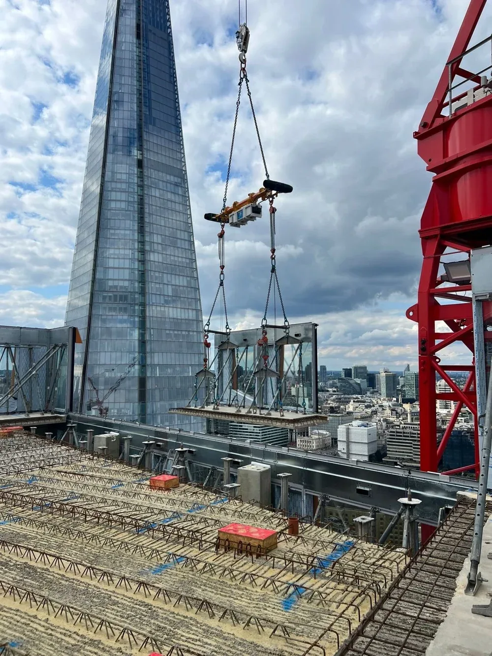 Construction site on the rooftop of a high-rise building in a city, with steel reinforcement bars and construction equipment, including a crane and safety barriers, beside the Shard skyscraper.