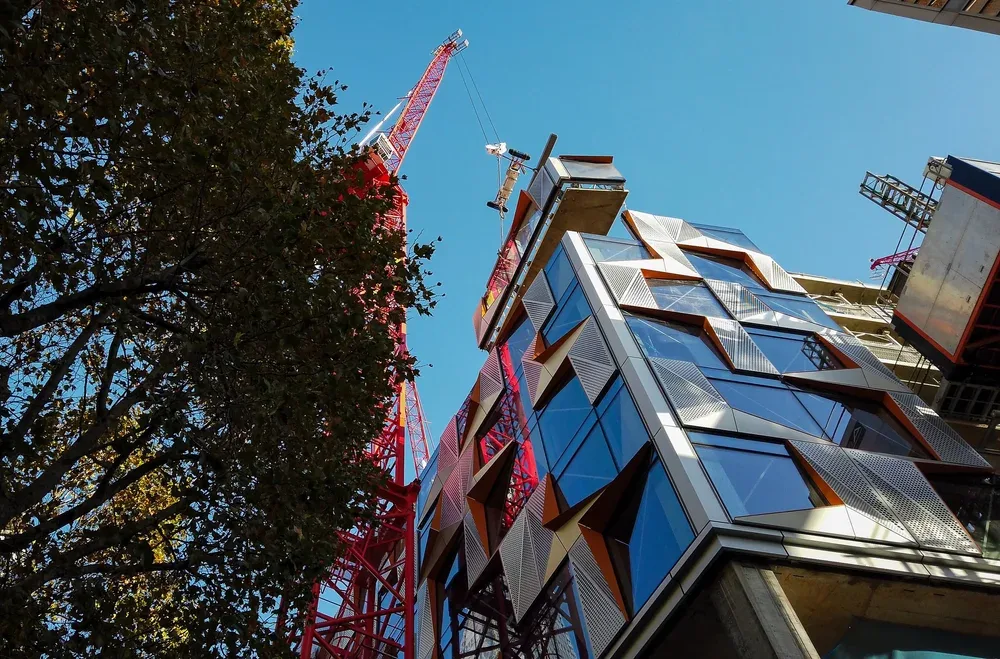 View of a modern building under construction with a red construction crane against a clear blue sky, and a tree on the left.