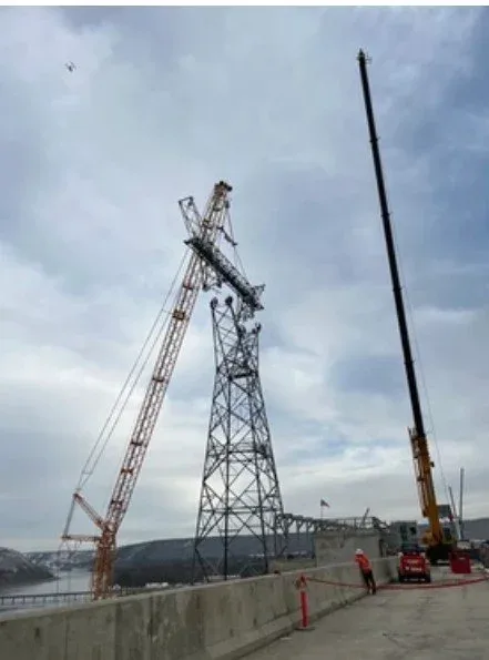 Construction workers using cranes to work on a tall metal power line tower along a waterfront.