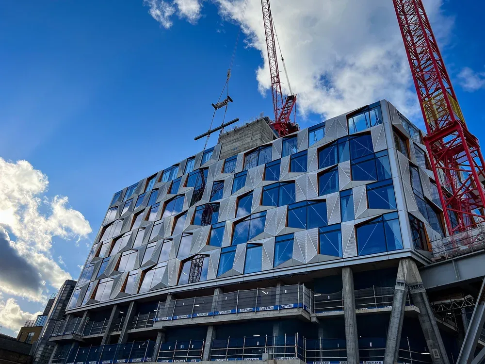 A modern building under construction with a geometric glass facade, a red construction crane, and a blue sky with scattered clouds.