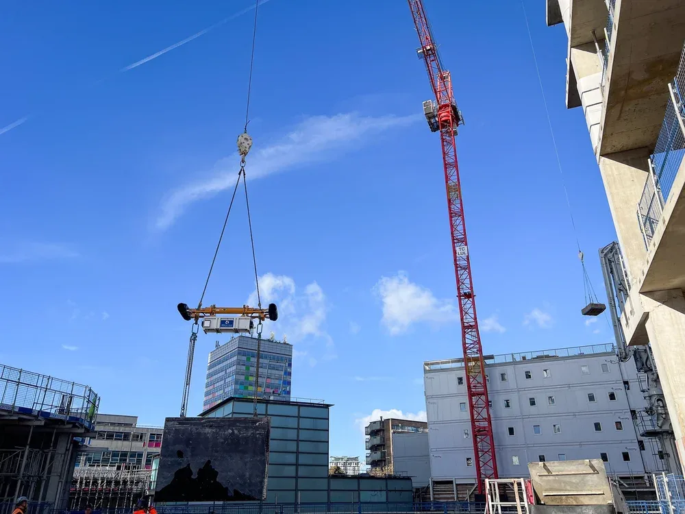 Construction site with a large red crane lifting a concrete slab, surrounded by buildings under construction against a blue sky with a few clouds.