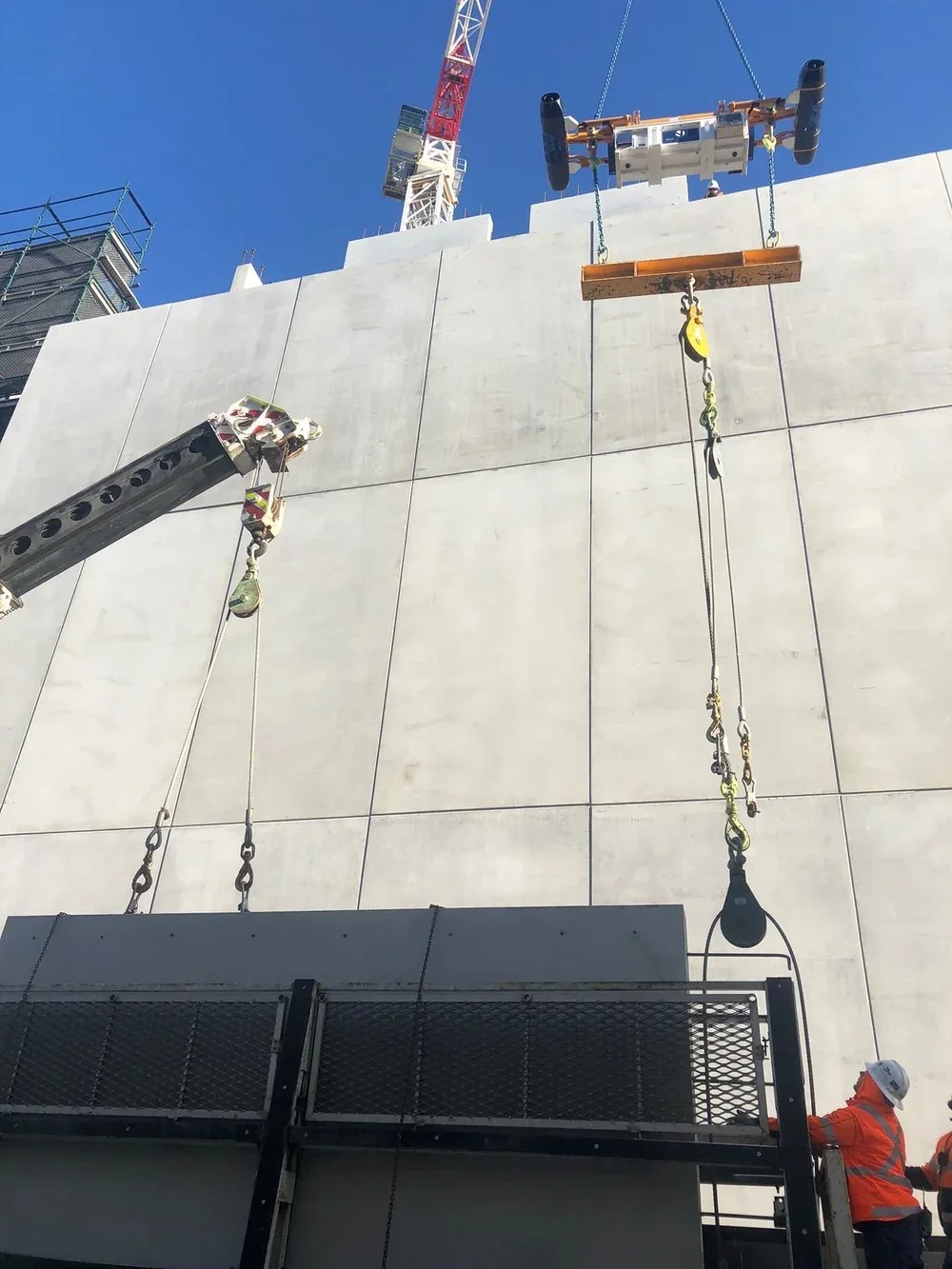 Construction workers in orange safety vests and white helmets working on a large white concrete wall at a construction site. A crane and construction equipment are visible above the wall.