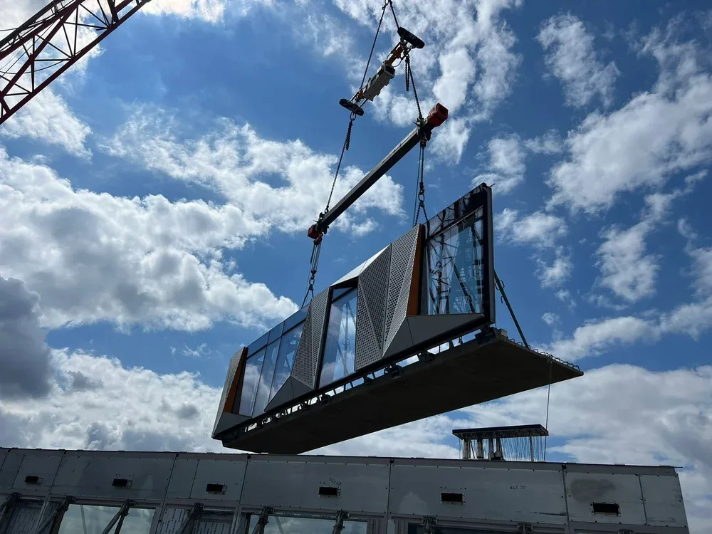 A building structure is being lifted by a crane and controlled by a VLN against a blue sky with white clouds.