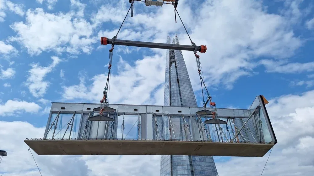 A glass platform being suspended in the air with a tall skyscraper in the background and blue sky with clouds above.