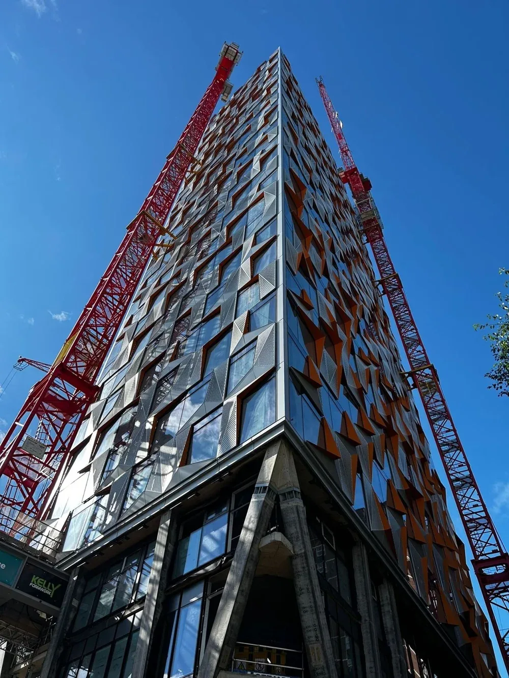 Looking up at a modern glass and metal high-rise building under construction with two red cranes on either side against a clear blue sky.