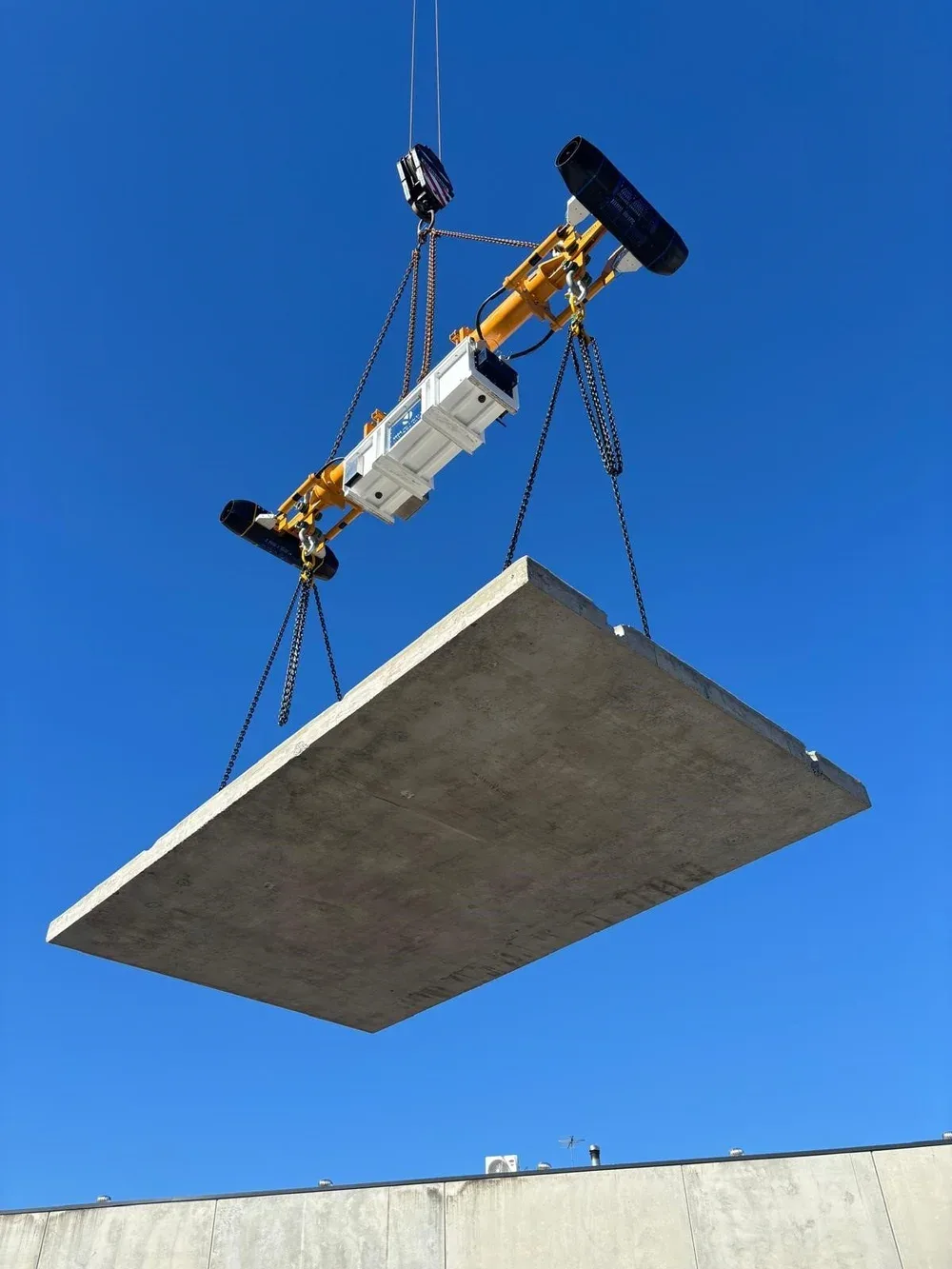 A large suspended concrete slab like a building floor or rooftop panel being hoisted by a construction crane against a clear blue sky.