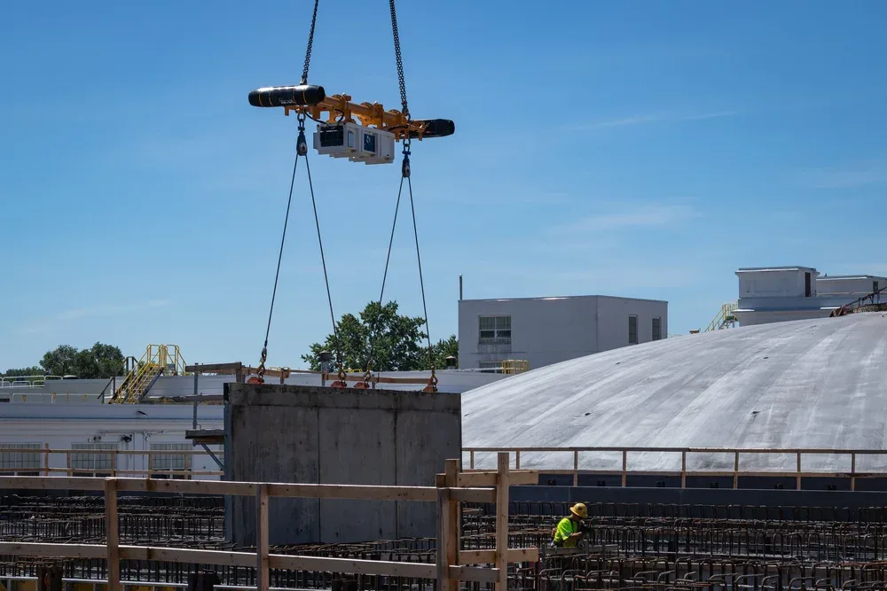 A worker wearing a yellow safety helmet and a reflective vest is working on a construction site with rebar and a large white domed roof, while a crane hoists a concrete slab.