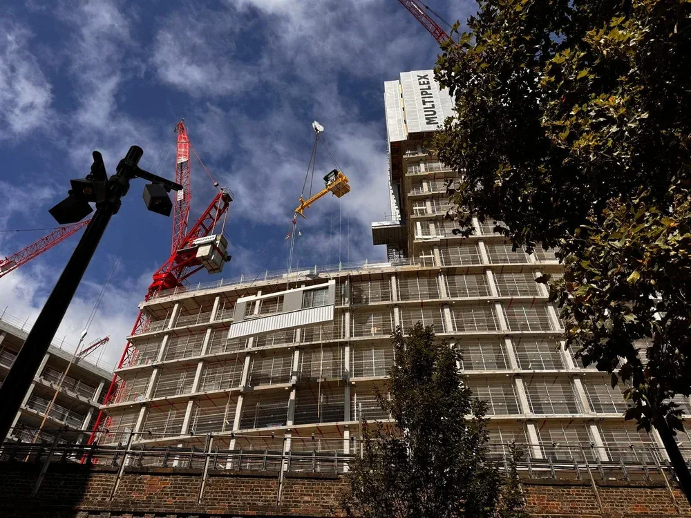 Construction site with a building under construction, cranes, a streetlight, and trees in the foreground.