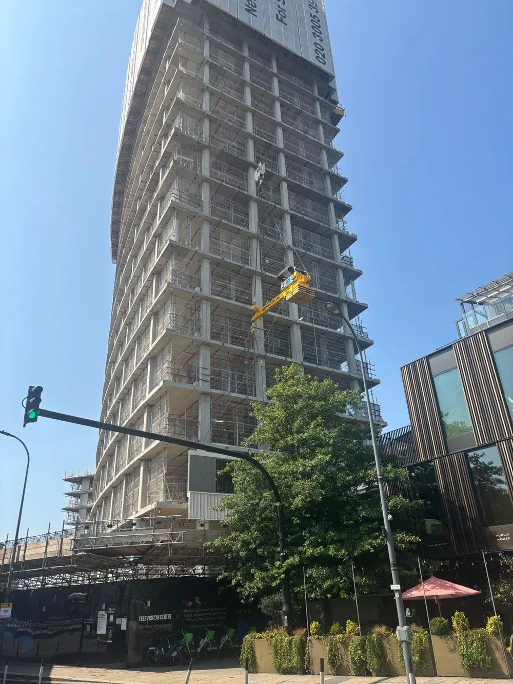 A tall high-rise building under construction with scaffolding and construction materials around the base. There is a green traffic light, a tree, and a modern building with glass windows in the foreground.