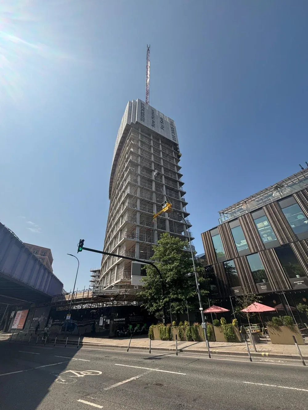A tall building under construction with scaffolding and a crane, next to a modern glass building with outdoor seating, in an urban area.