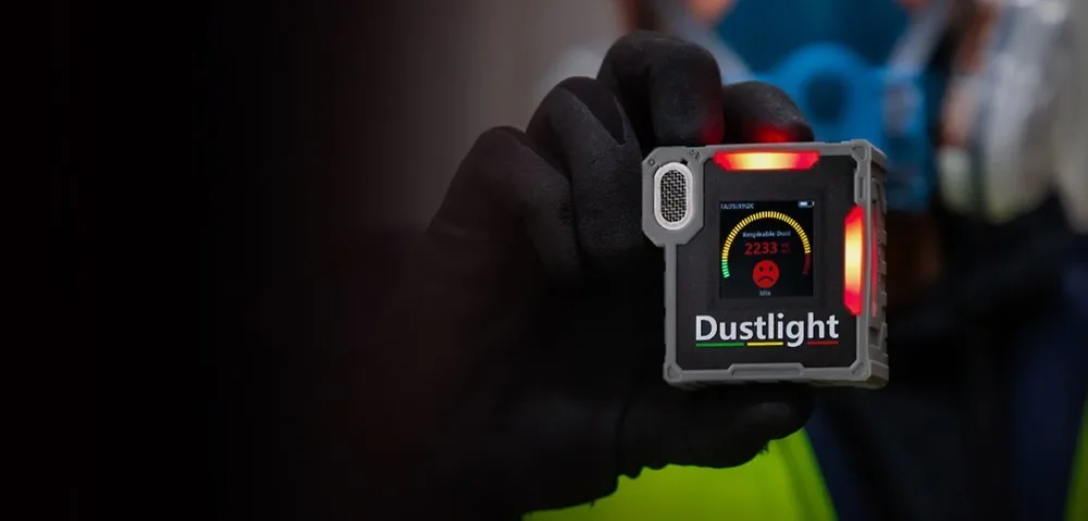 A person wearing black gloves holding a Dustlight device that displays dust pollution levels with a red warning indicator.